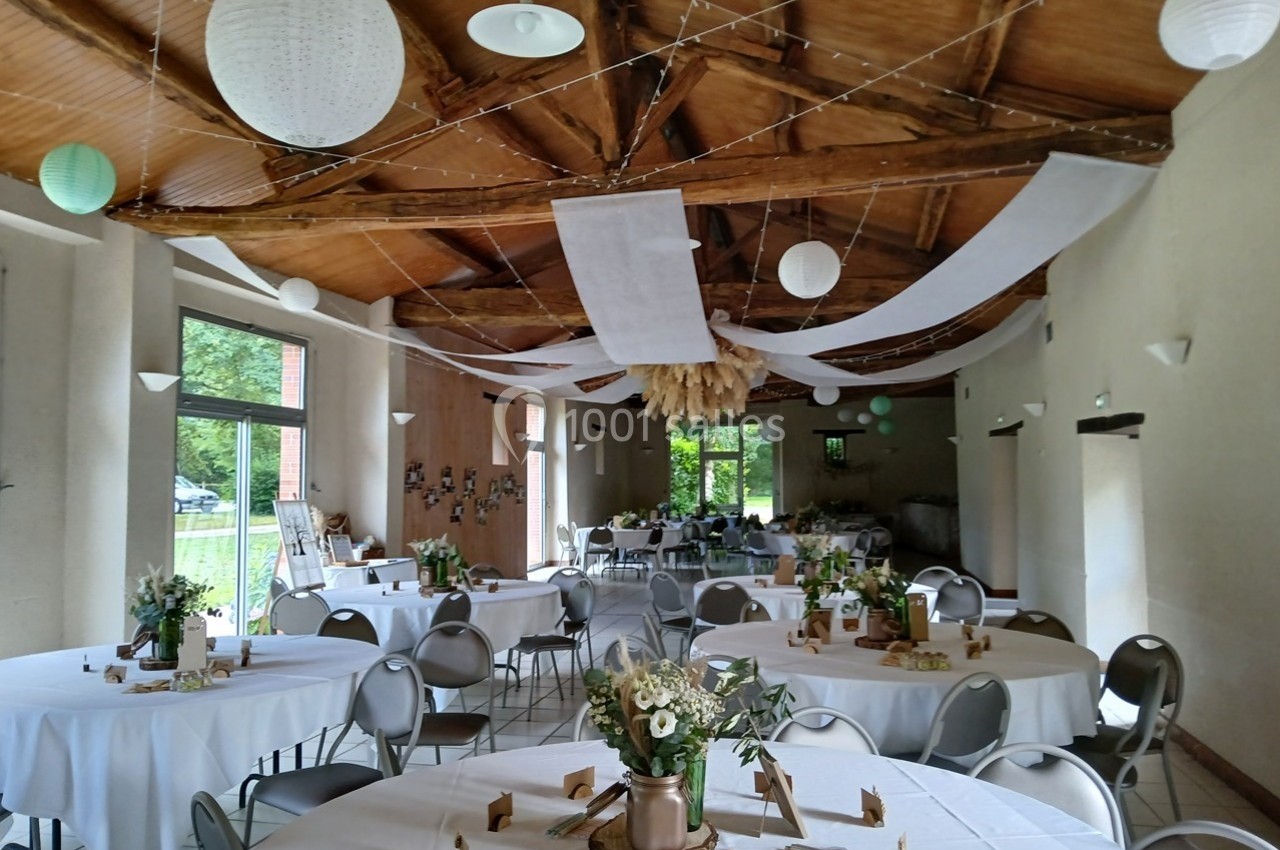 Salle décorée pour un événement avec des tables rondes dressées, des fleurs et des guirlandes suspendues au plafond en bois.