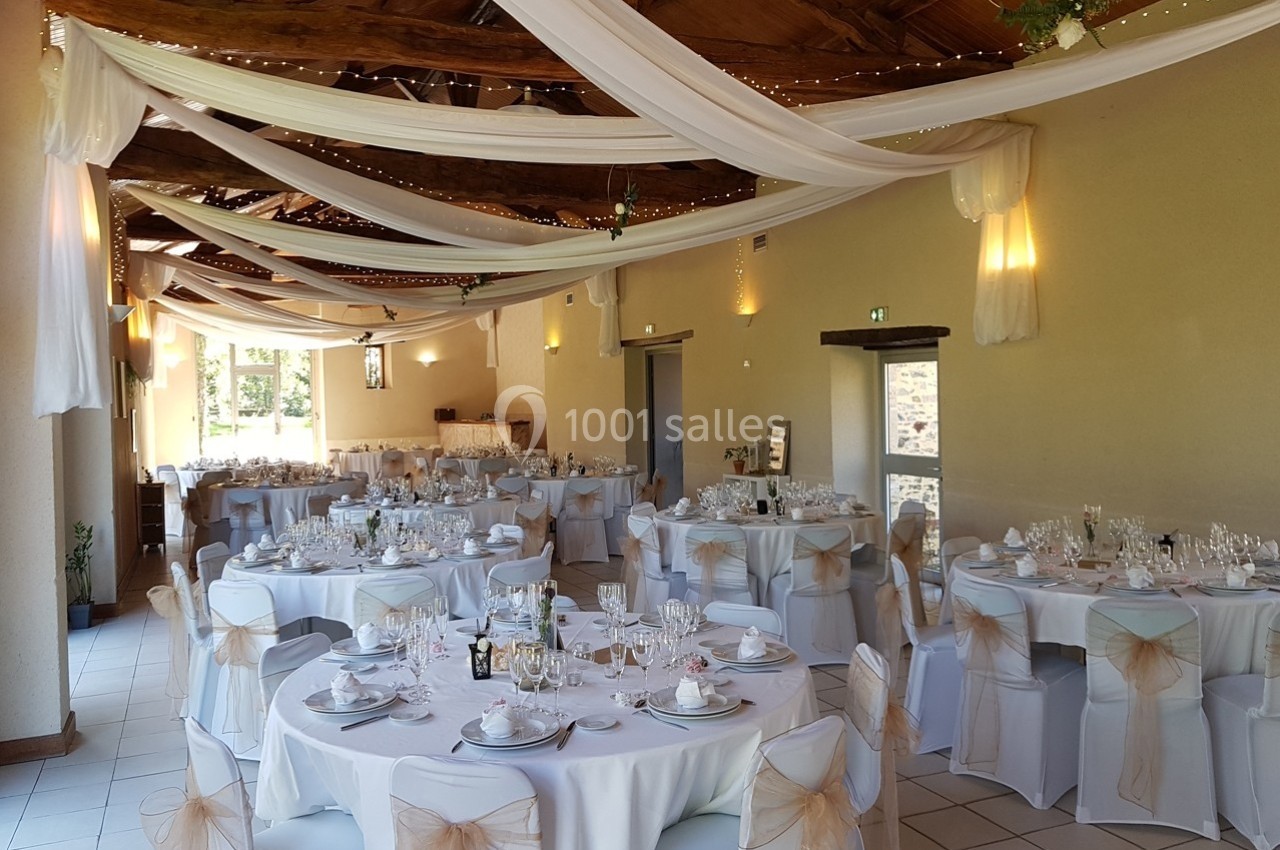 Salle de réception décorée avec des nappes blanches, des rubans beige sur les chaises et des drapés suspendus au plafond.