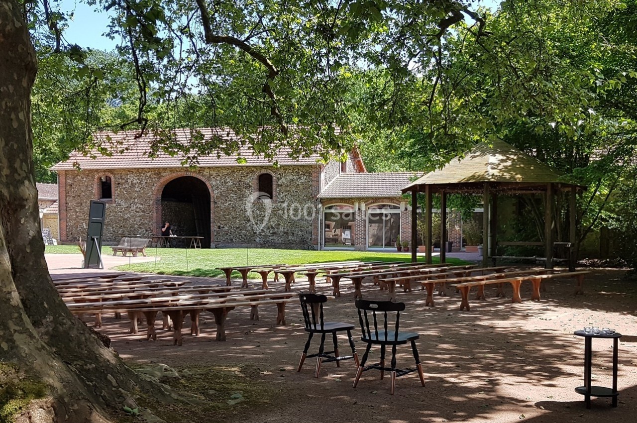 Cour extérieure avec bancs en bois, chaises noires, arbres, et bâtiments en briques sous un ciel dégagé.