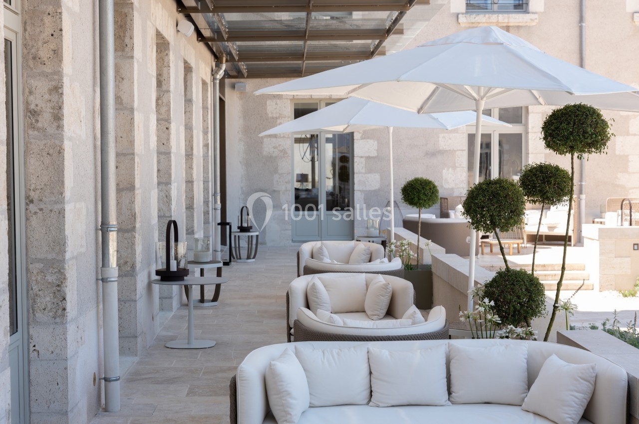 Terrasse élégante avec mobilier beige, parasols blancs et plantes taillées, adossée à un bâtiment en pierre claire.