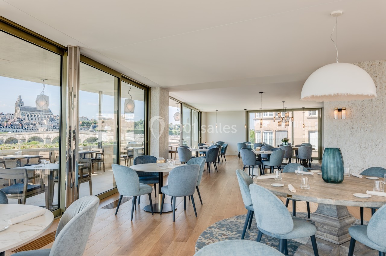 Salle de restaurant lumineuse avec grandes baies vitrées offrant une vue sur un pont et des bâtiments historiques.