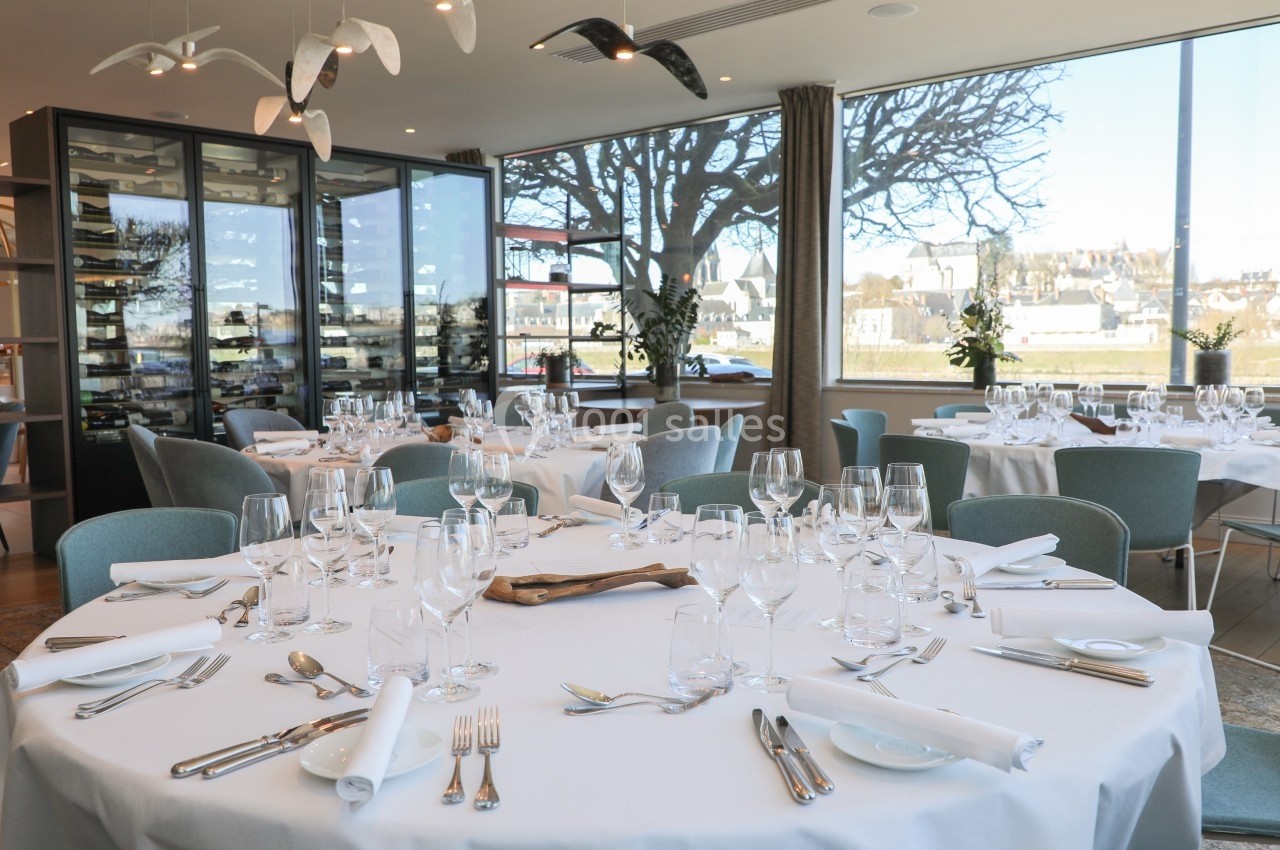 Salle de restaurant lumineuse avec tables rondes dressées, grandes baies vitrées et vue sur un paysage urbain.