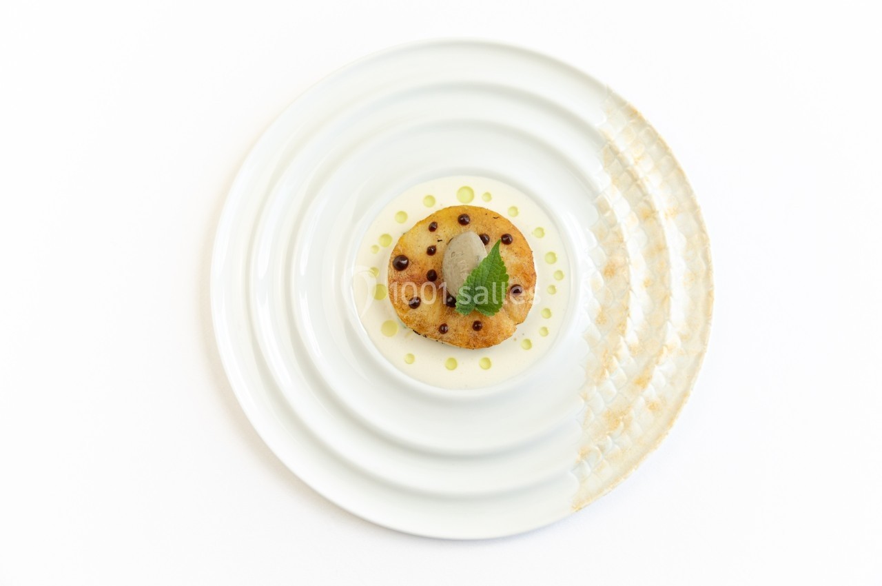 Assiette blanche avec un plat gastronomique composé d'un biscuit rond, d'une crème, d'une feuille verte et de décorations.