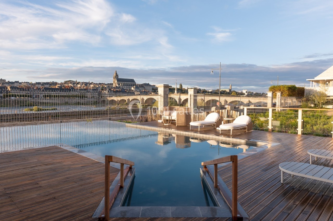 Piscine extérieure avec vue sur un pont en pierre, une rivière et une ville historique sous un ciel partiellement nuageux.