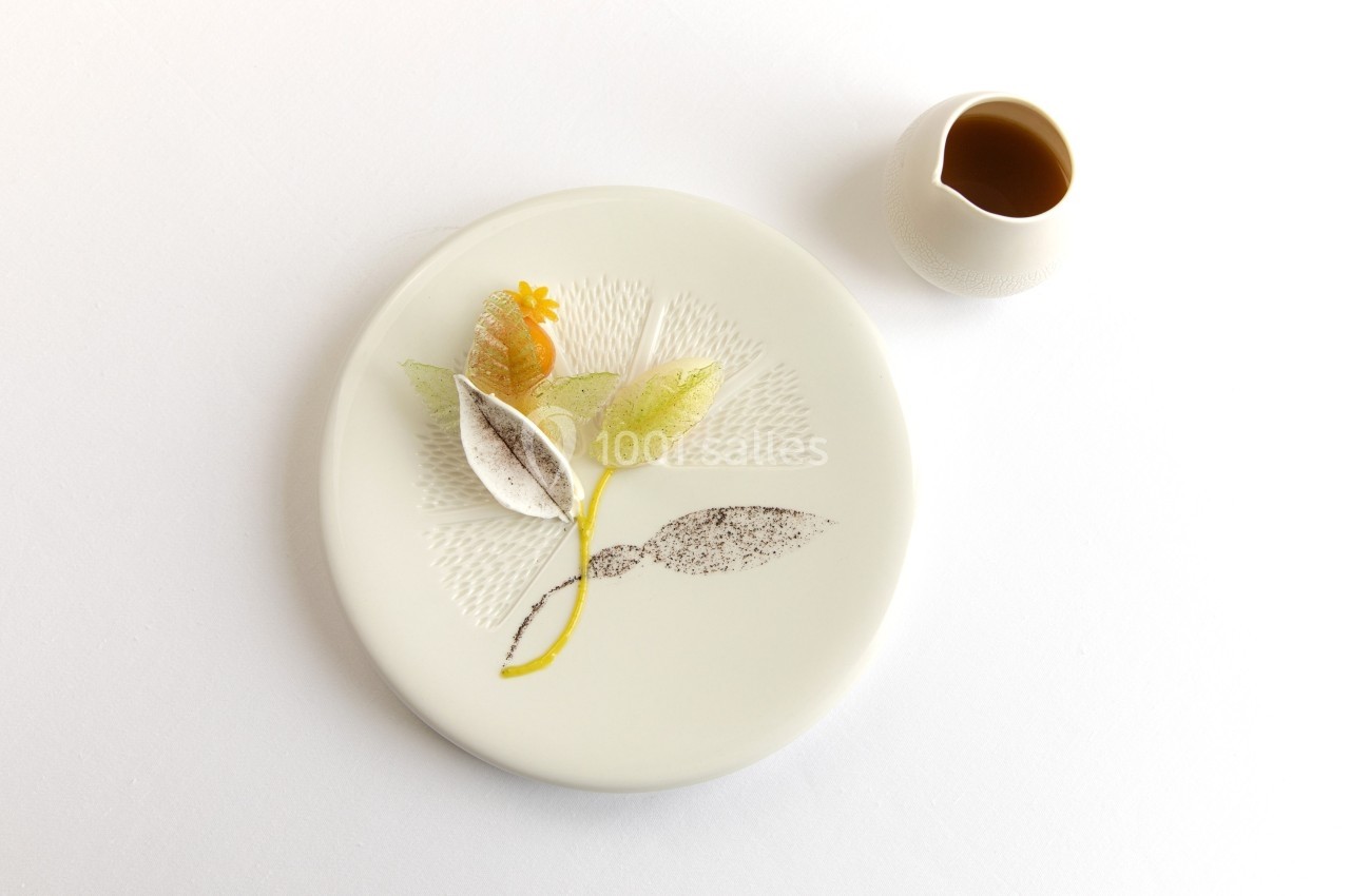 Assiette minimaliste décorée de feuilles comestibles et d'une fleur, accompagnée d'une petite carafe de sauce.
