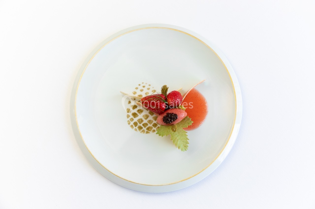 Assiette blanche contenant une présentation soignée de fraises, feuilles et sauce rouge sur un motif doré.