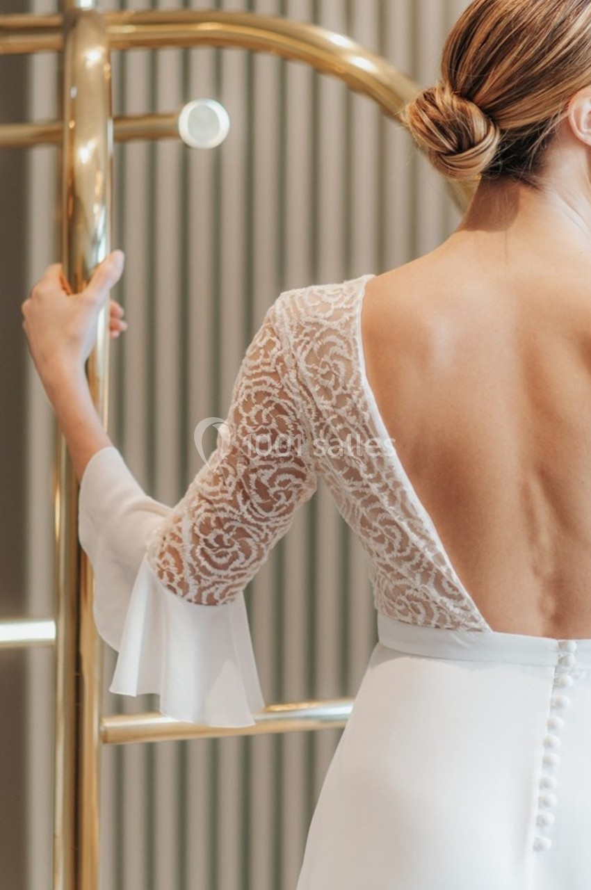 Femme en robe blanche au dos ouvert avec manches en dentelle, tenant une barre dorée dans un intérieur lumineux.