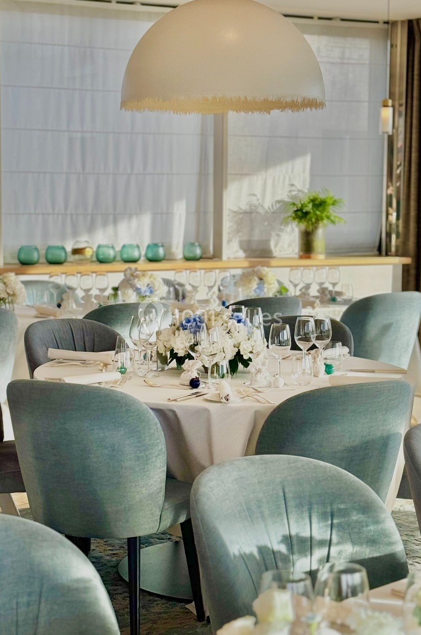 Salle élégante avec tables rondes dressées, chaises en velours gris-bleu et décoration florale blanche et bleue.