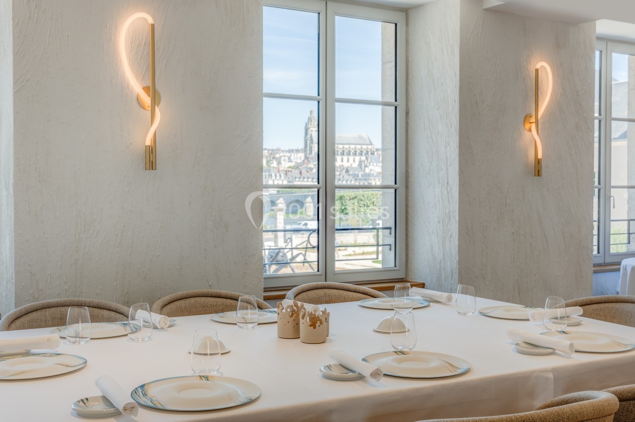 Salle lumineuse avec tables dressées, grandes fenêtres offrant une vue sur des bâtiments historiques et des appliques…