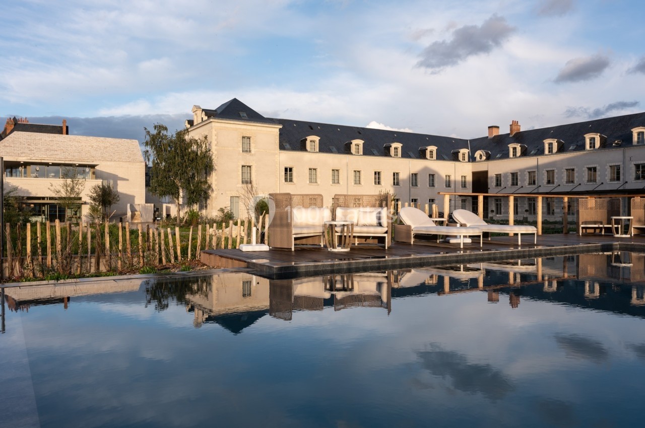 Bâtiment en pierre avec toit en ardoise, terrasse aménagée et piscine reflétant le ciel partiellement nuageux.