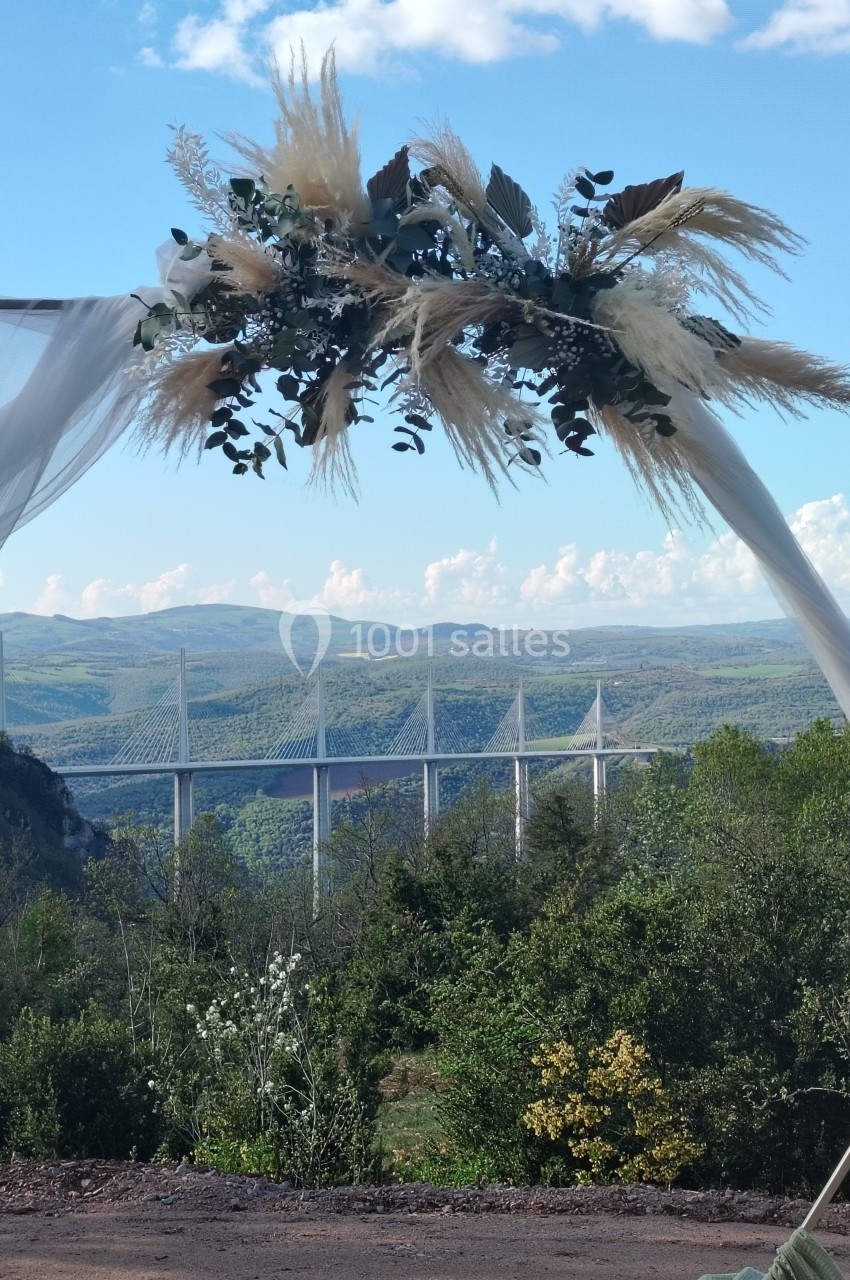 Vue sur le viaduc de Millau encadrée par une arche décorée de feuillages et de pampas, sous un ciel dégagé.