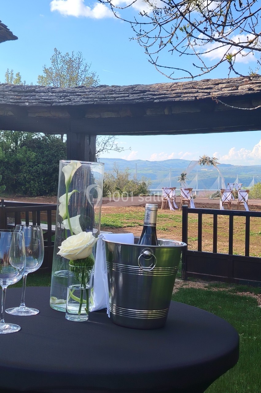 Table dressée en extérieur avec des verres, un seau à champagne et des fleurs, vue sur un paysage champêtre.