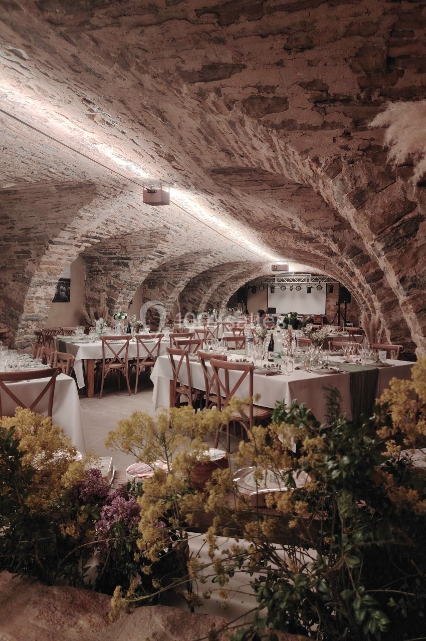 Salle voûtée en pierre aménagée en restaurant, avec tables dressées et décorations florales au premier plan.