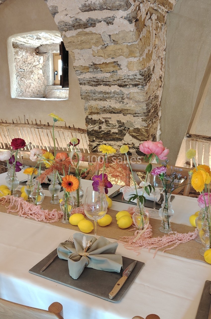 Table décorée avec des fleurs colorées, des citrons et des nappes naturelles dans une salle en pierre lumineuse.