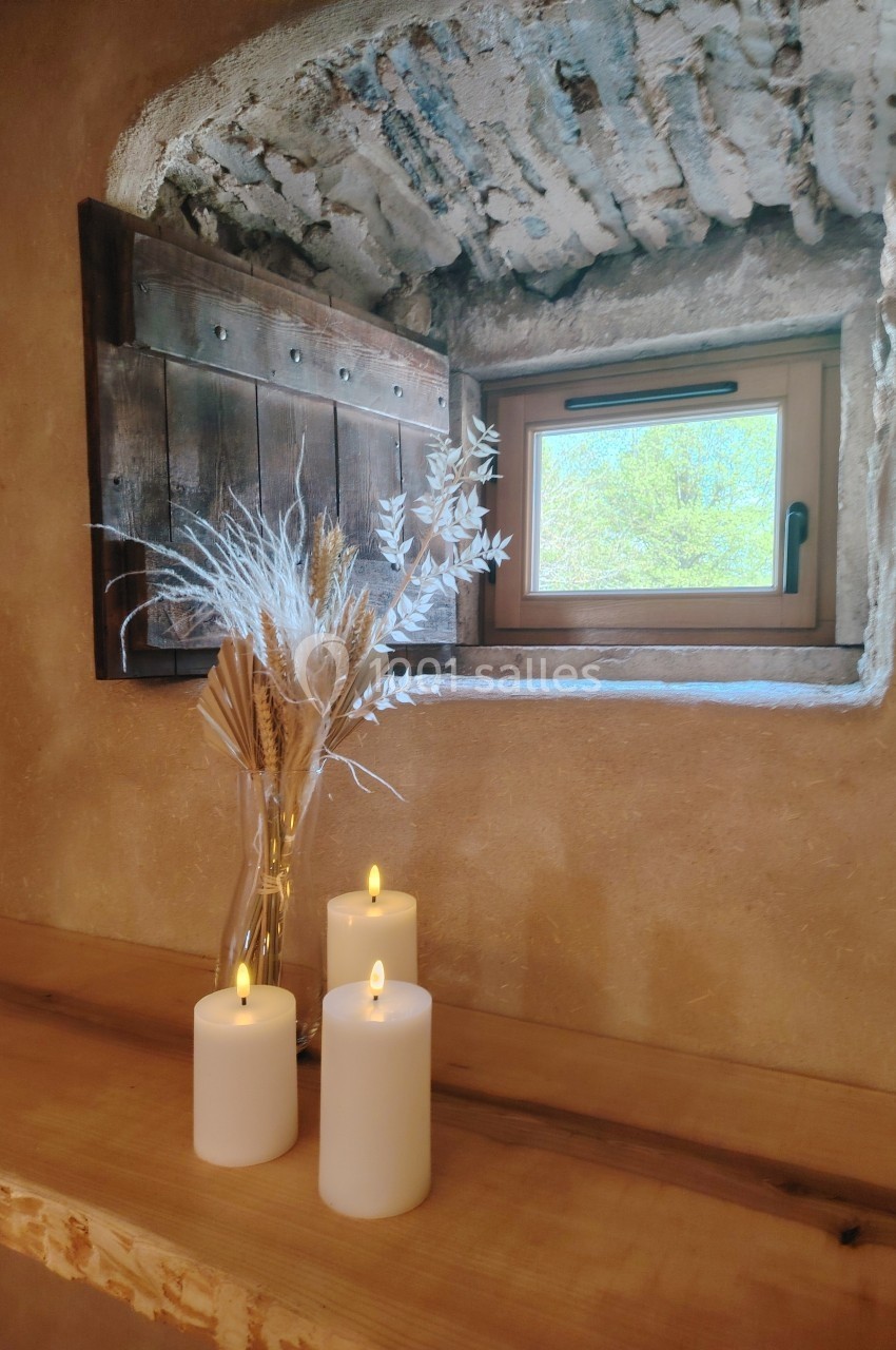 Bougies allumées et vase de fleurs séchées posés sur une étagère en bois devant une fenêtre rustique.