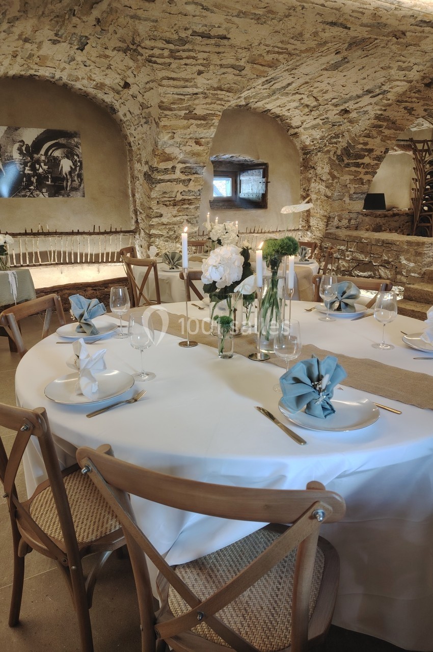Salle voûtée en pierre avec une table ronde dressée, ornée de fleurs blanches et serviettes pliées bleues.