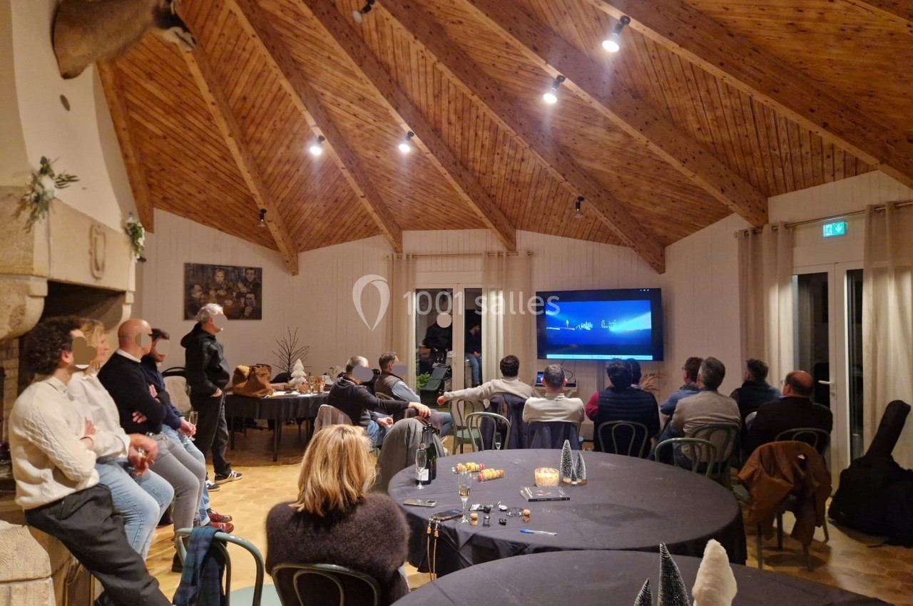 Groupe de personnes assises et debout dans une salle avec plafond en bois, regardant un écran diffusant une vidéo.