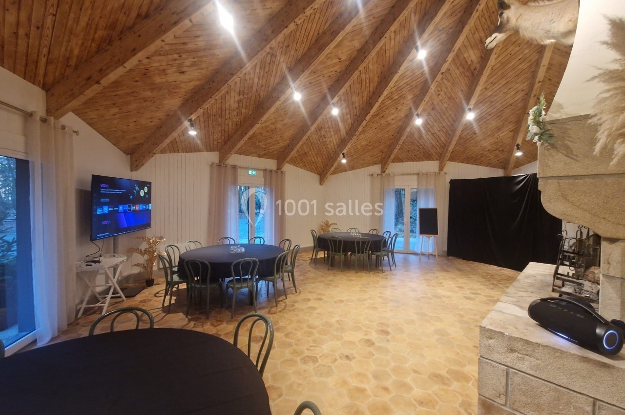 Salle lumineuse avec plafond en bois, tables rondes noires, cheminée en pierre et écran mural.