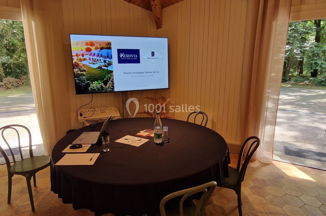 Salle lumineuse avec une table ronde, des chaises, un écran affichant une présentation et une vue sur un jardin boisé.