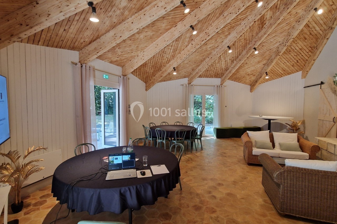 Salle lumineuse avec plafond en bois, tables rondes, canapé en osier, cheminée et écran de présentation.
