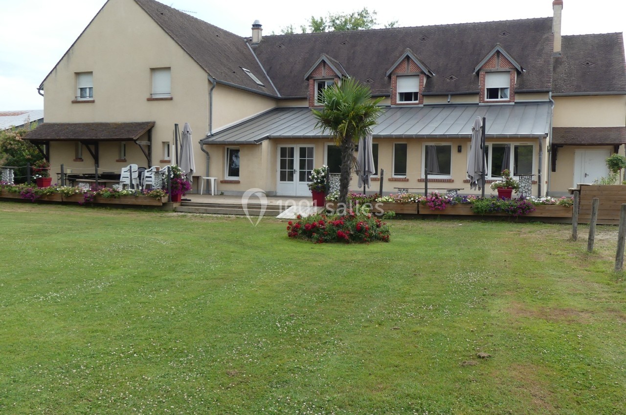 Grande maison avec terrasse fleurie, parasols et pelouse bien entretenue, entourée d'un environnement verdoyant.