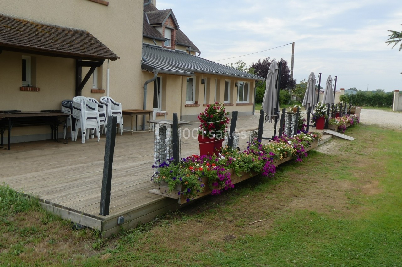 Terrasse en bois avec chaises empilées, parasols fermés et jardinières fleuries, bordée d'une pelouse.