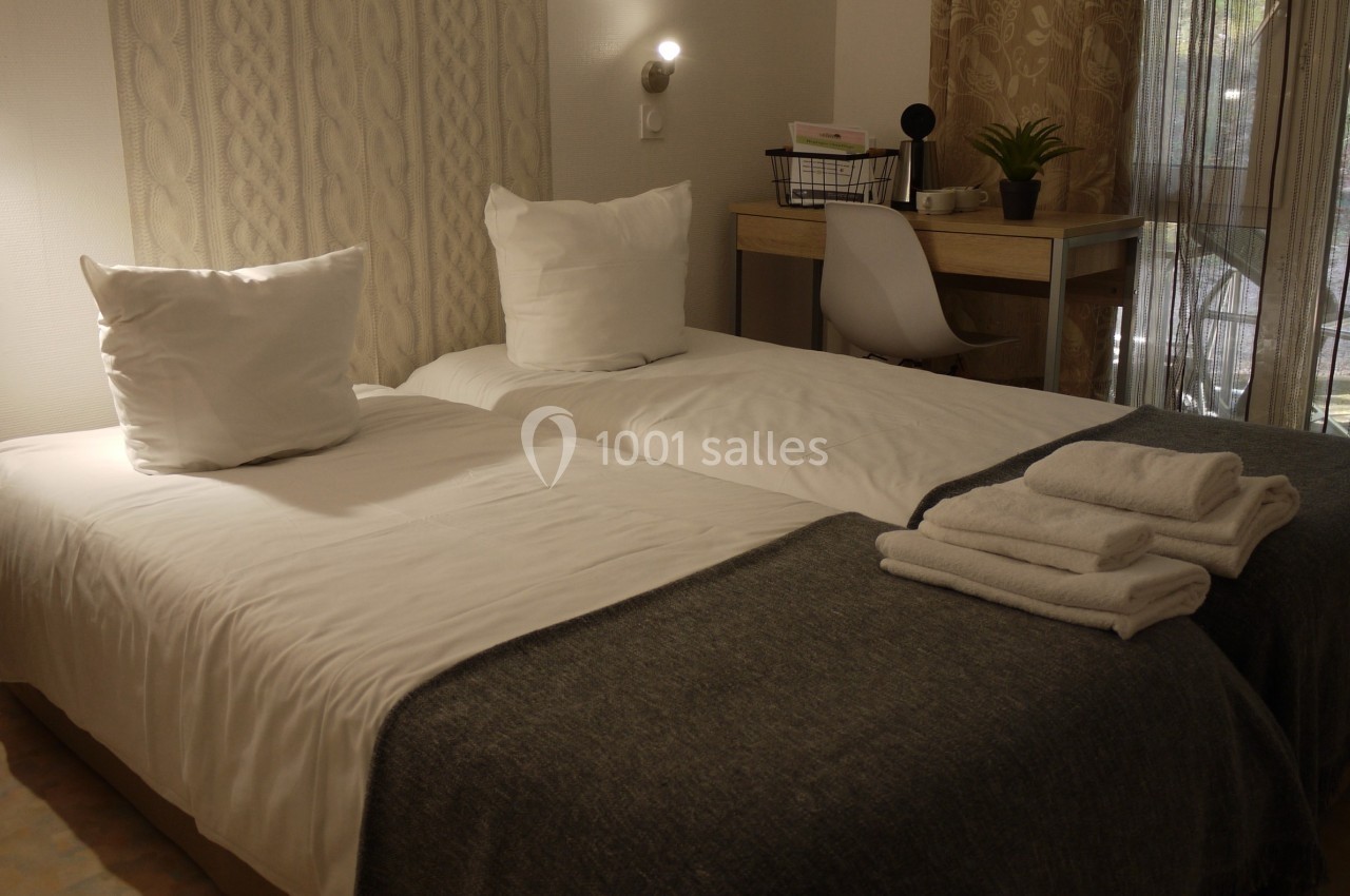 Deux lits simples avec draps blancs, table de travail, chaise et serviettes pliées dans une chambre lumineuse.