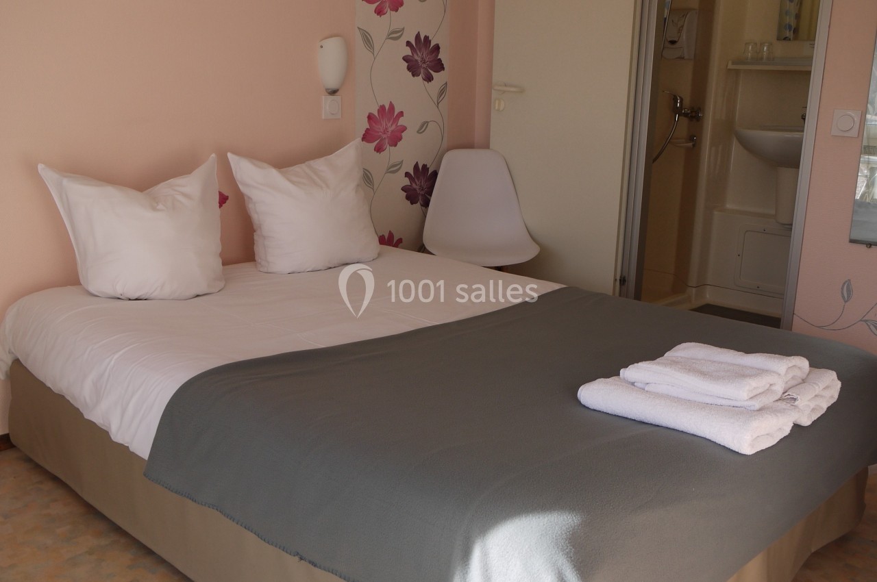 Chambre avec lit double, draps blancs, couverture grise, serviettes pliées, mur décoré de motifs floraux.