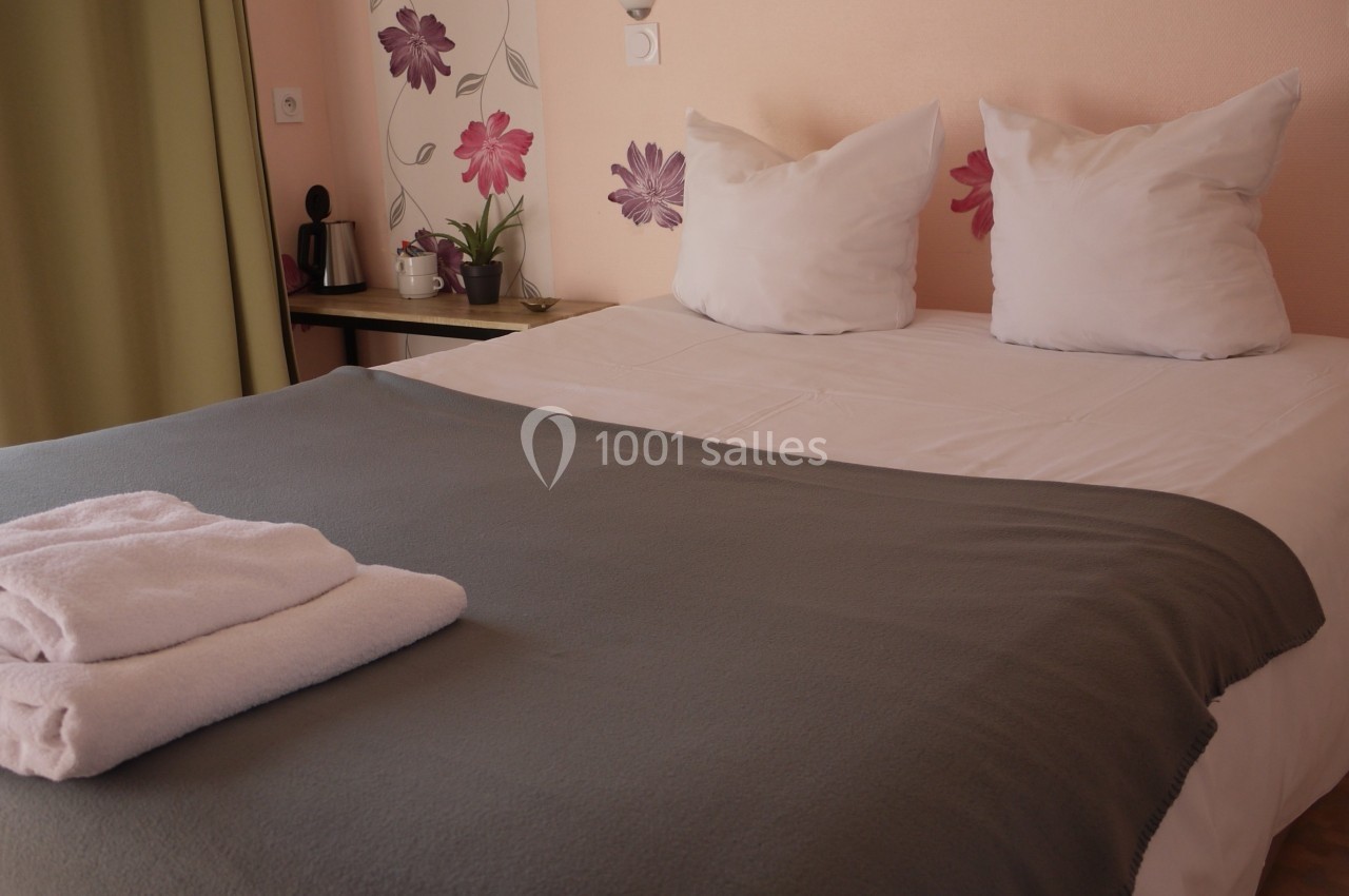 Lit double avec couverture grise, oreillers blancs, serviettes pliées au premier plan et décoration florale murale.