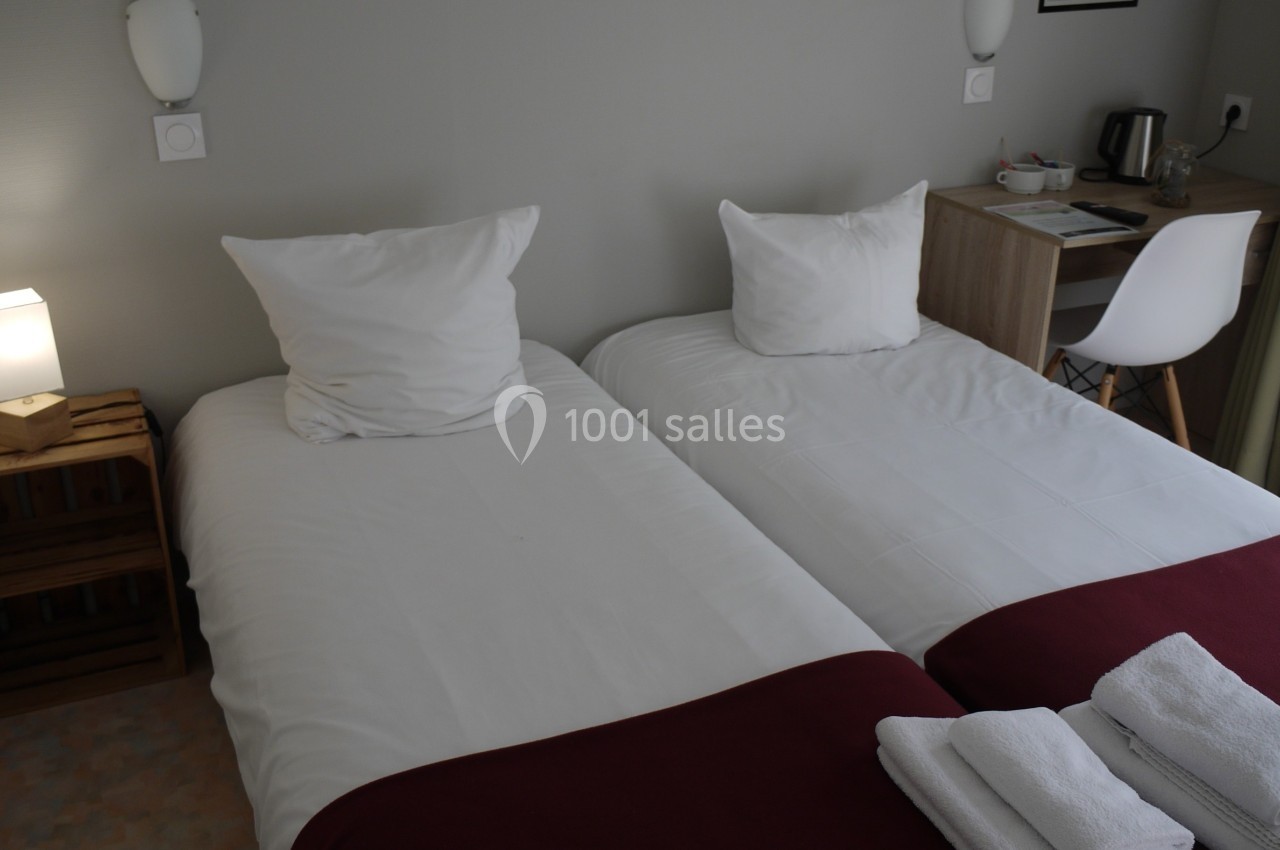 Deux lits simples avec draps blancs et couvertures rouges, une table de chevet, un bureau et une chaise dans une chambre.