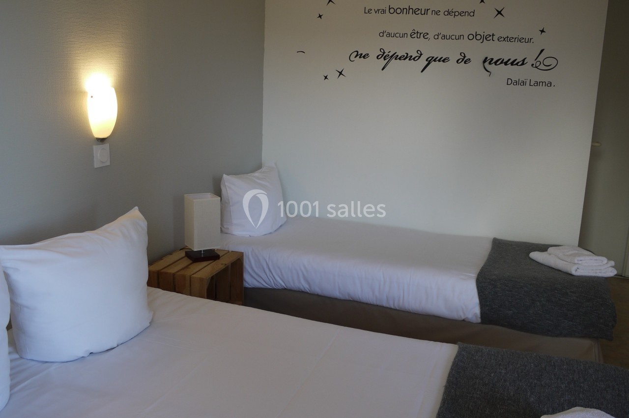 Deux lits simples avec linge blanc et plaid gris dans une chambre sobre, éclairée par une applique murale.