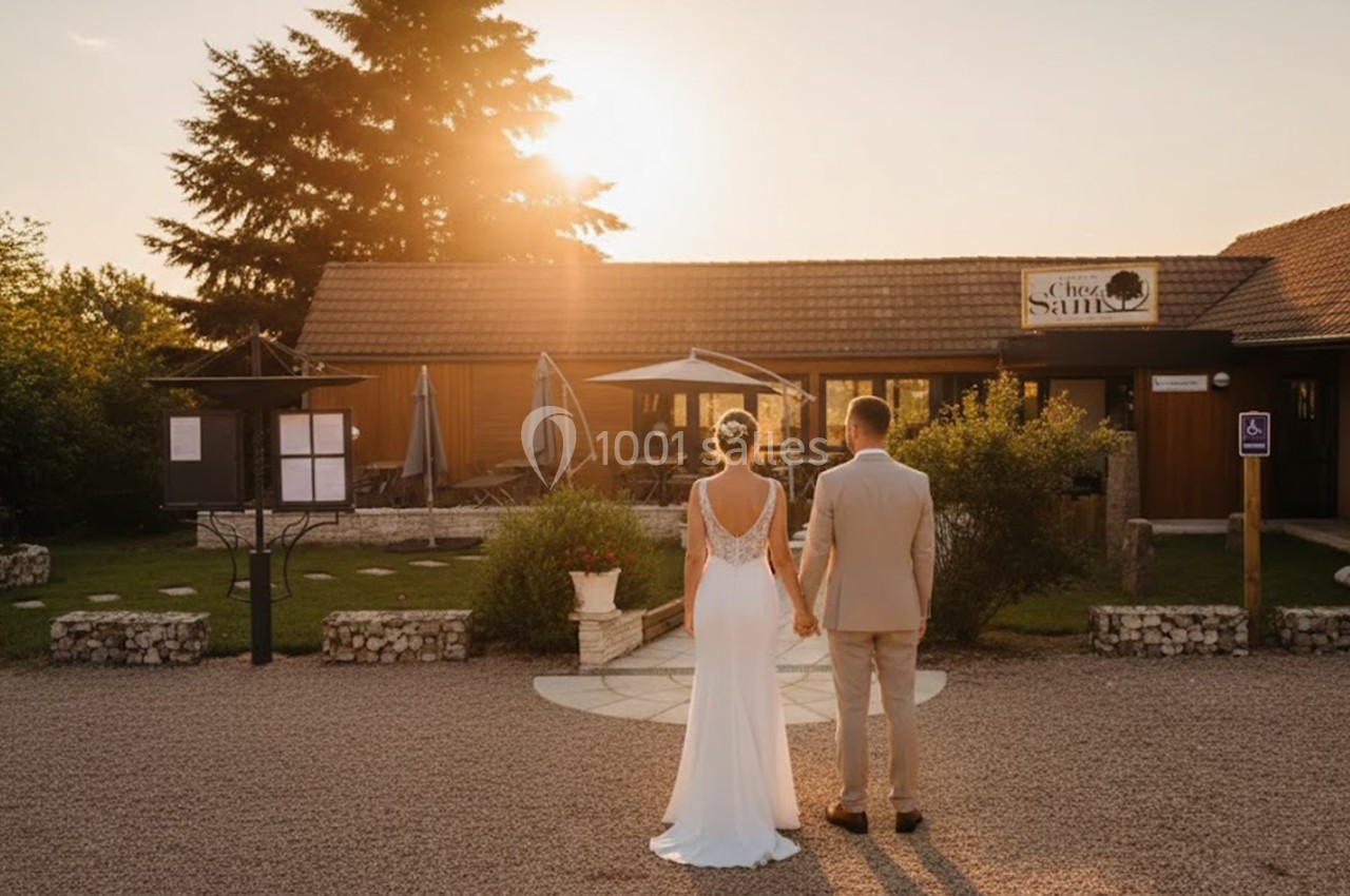 Un couple de dos en tenue de mariage regarde un bâtiment en bois au coucher du soleil.