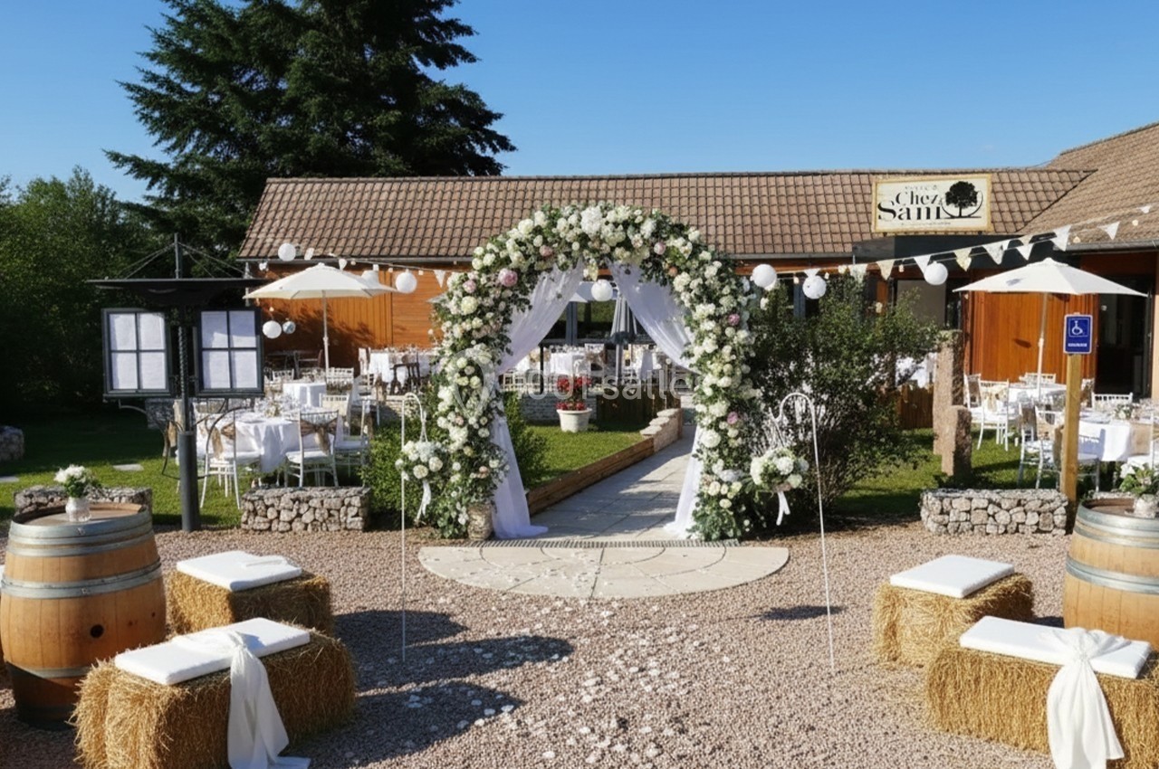 Arche fleurie devant une salle de réception en extérieur, entourée de tables décorées et de bottes de paille.