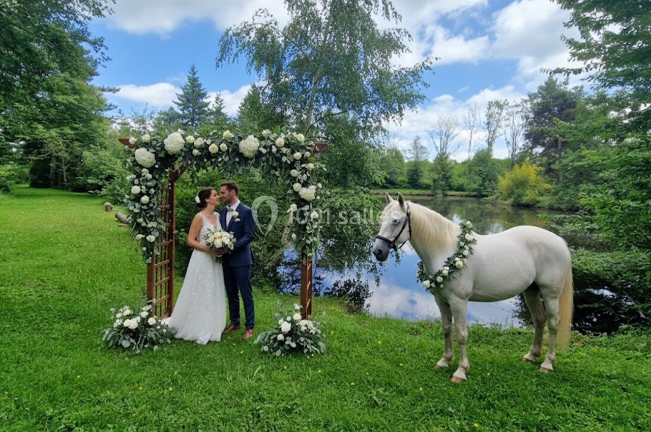 Un couple en tenue de mariage se tient sous une arche fleurie près d'un cheval blanc décoré, devant un étang.