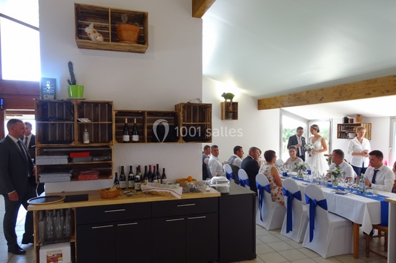 Salle de réception avec invités assis à des tables décorées, buffet avec bouteilles de vin et étagères en bois au mur.