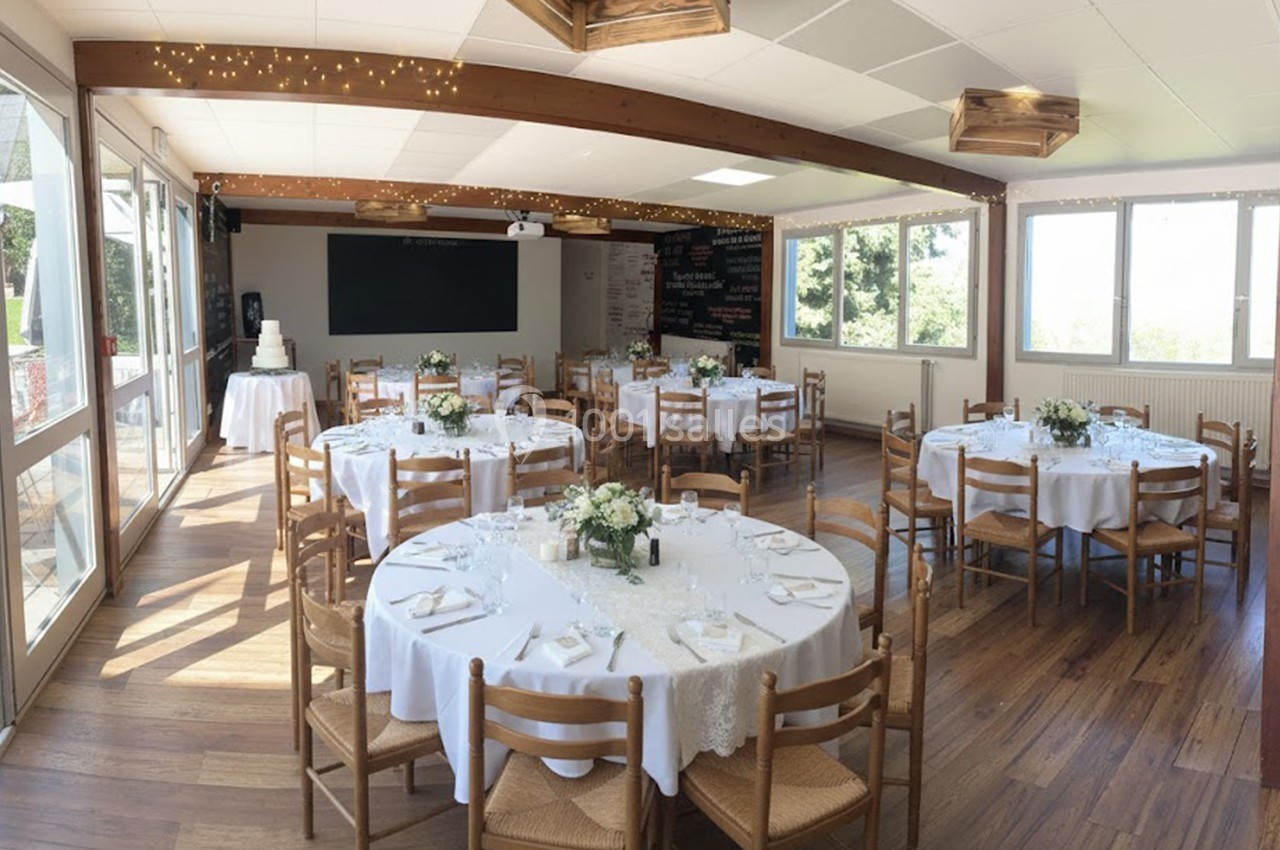 Salle lumineuse avec tables rondes dressées pour un événement, décorée de fleurs blanches et éclairée par des guirlandes.