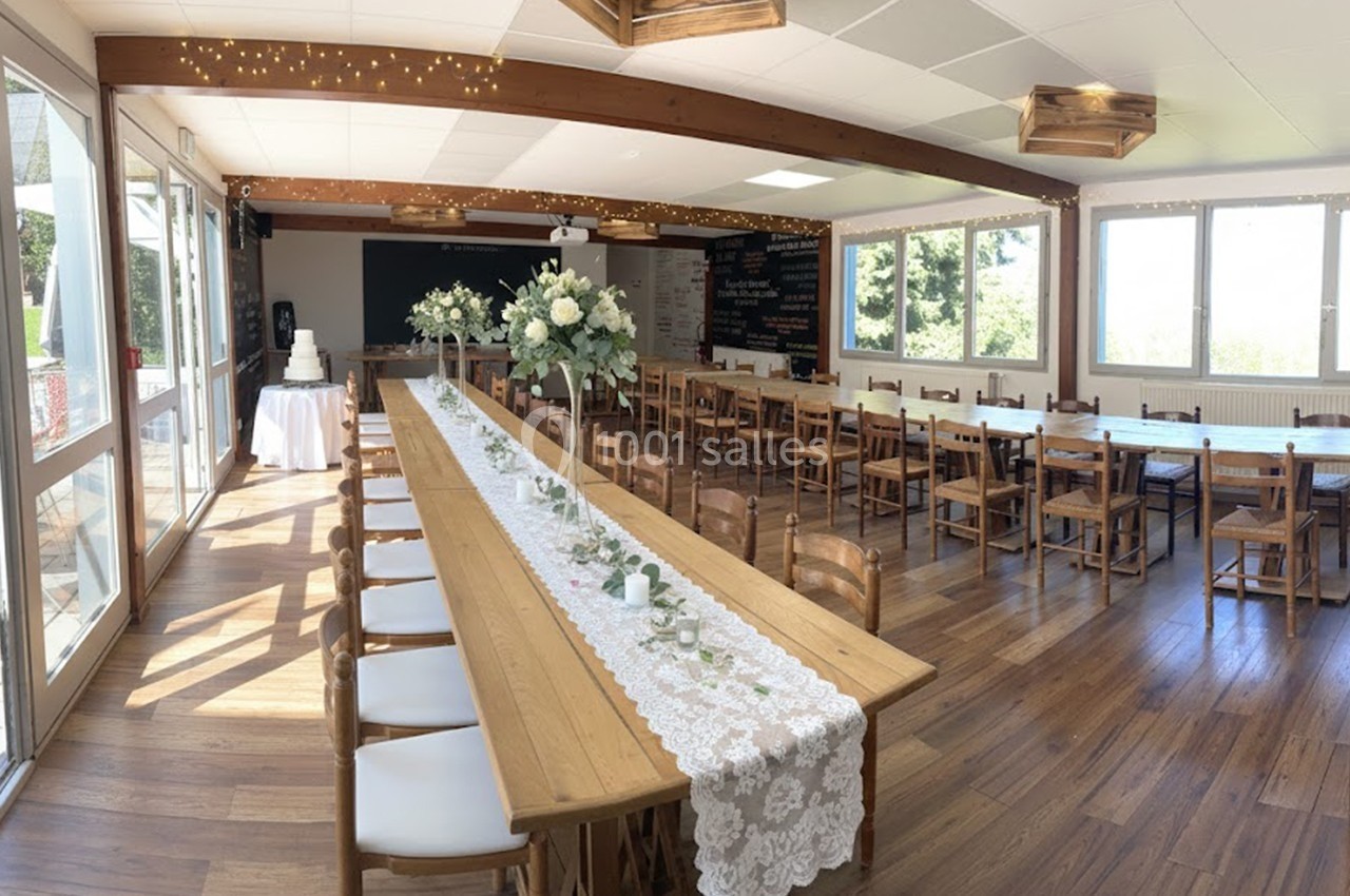 Salle de réception lumineuse avec une longue table décorée de fleurs et dentelle, entourée de chaises en bois.