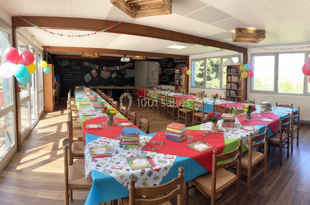 Salle décorée pour une fête d'anniversaire avec tables alignées, nappes colorées, ballons et livres en centre de table.