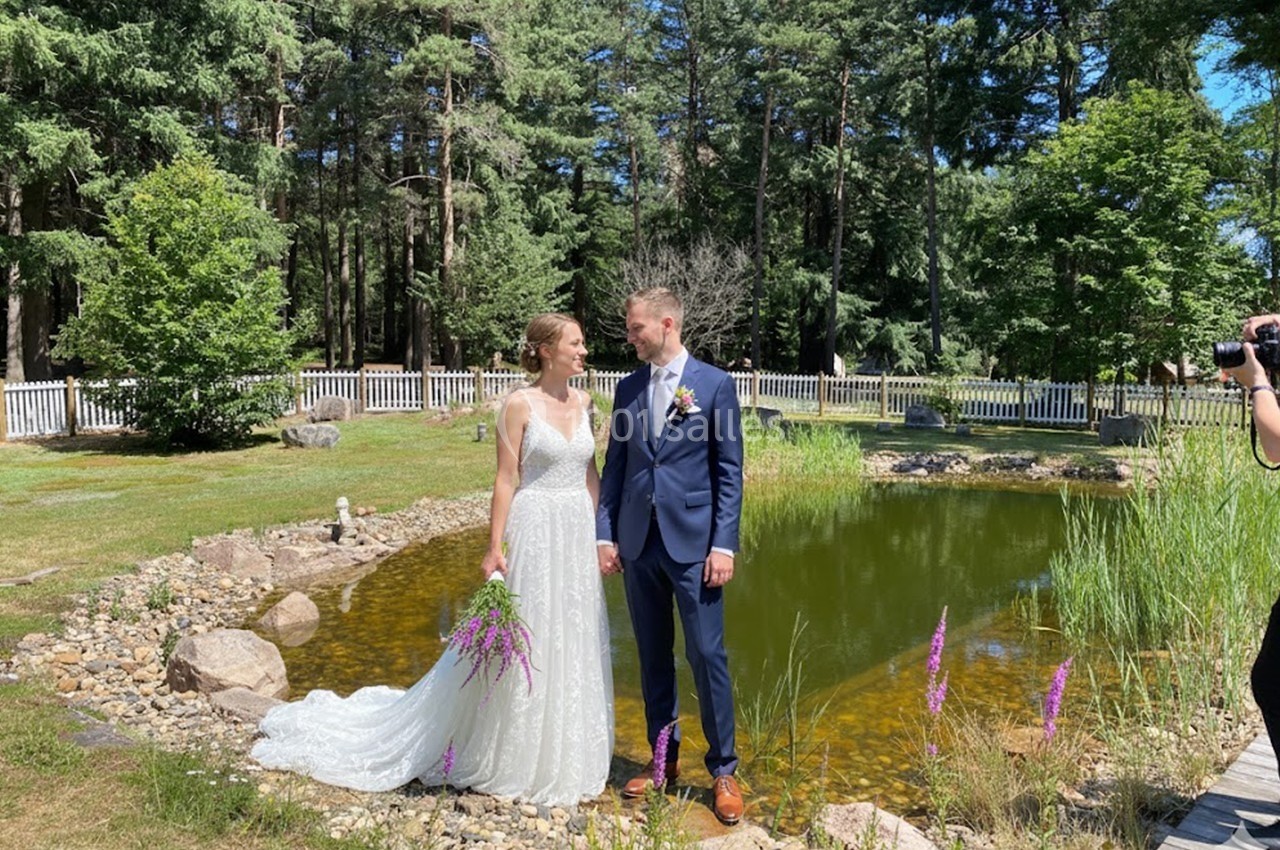 Un couple en tenue de mariage pose devant un petit étang entouré de verdure et de fleurs.