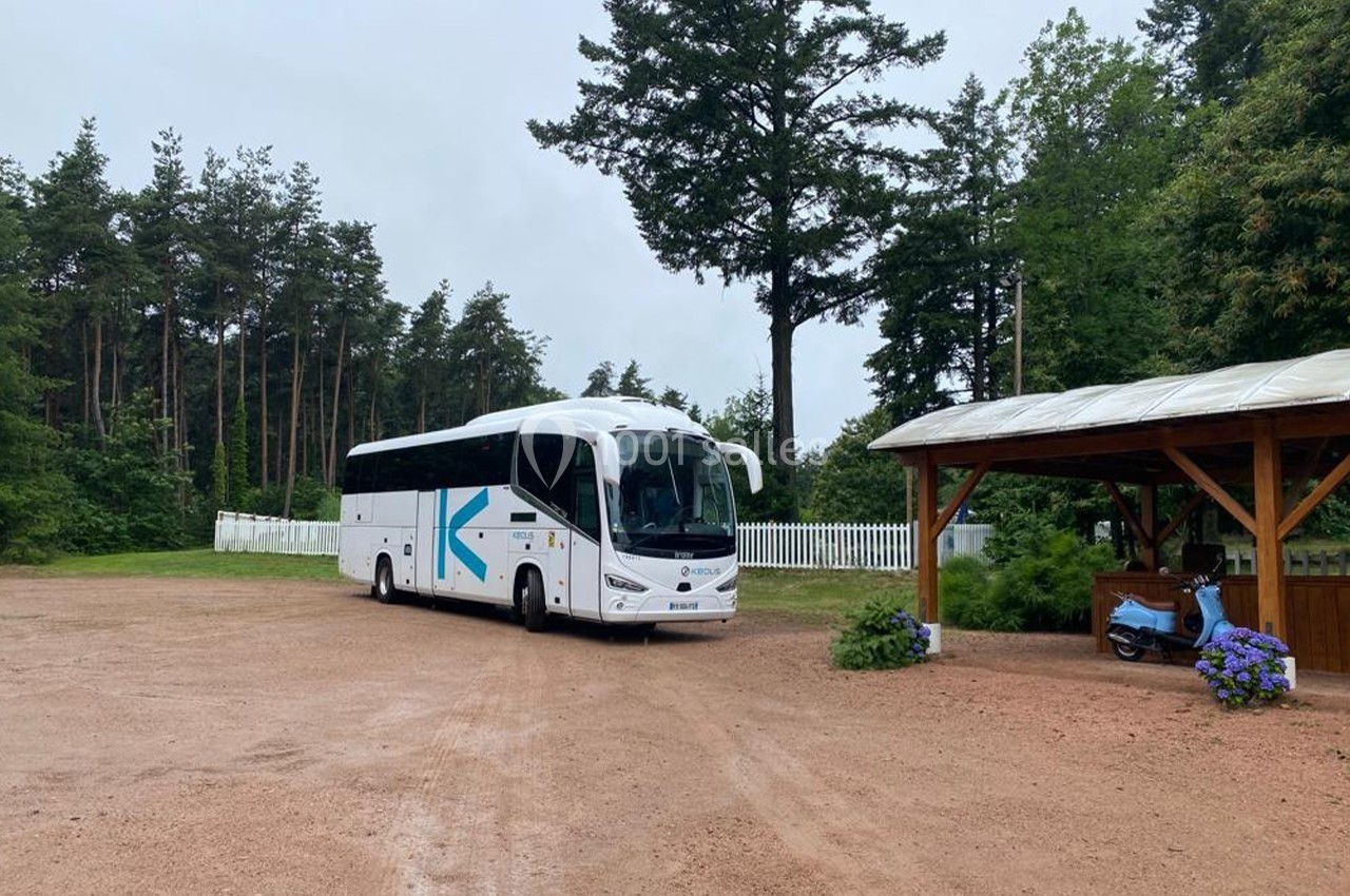 Autocar blanc stationné sur un terrain en gravier, entouré d'arbres et d'une petite structure en bois.