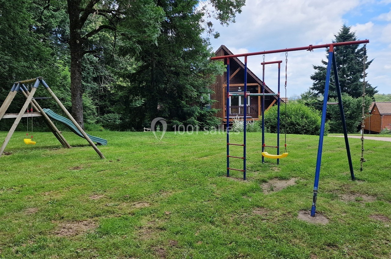 Aire de jeux extérieure avec balançoire, toboggan et échelle, située sur une pelouse près d'une maison en bois.