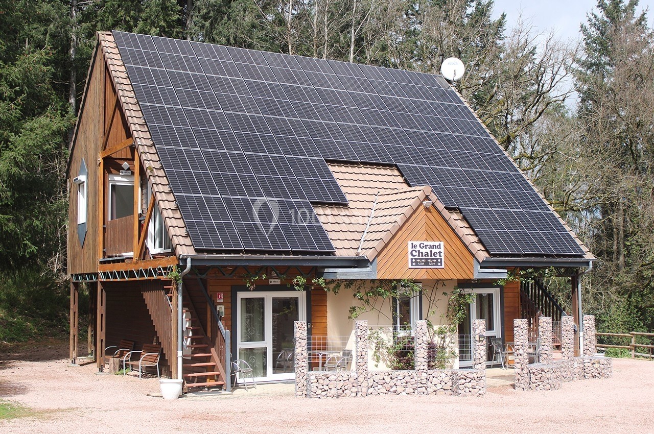 Chalet en bois avec toit incliné recouvert de panneaux solaires, entouré de végétation et d'une allée en gravier.