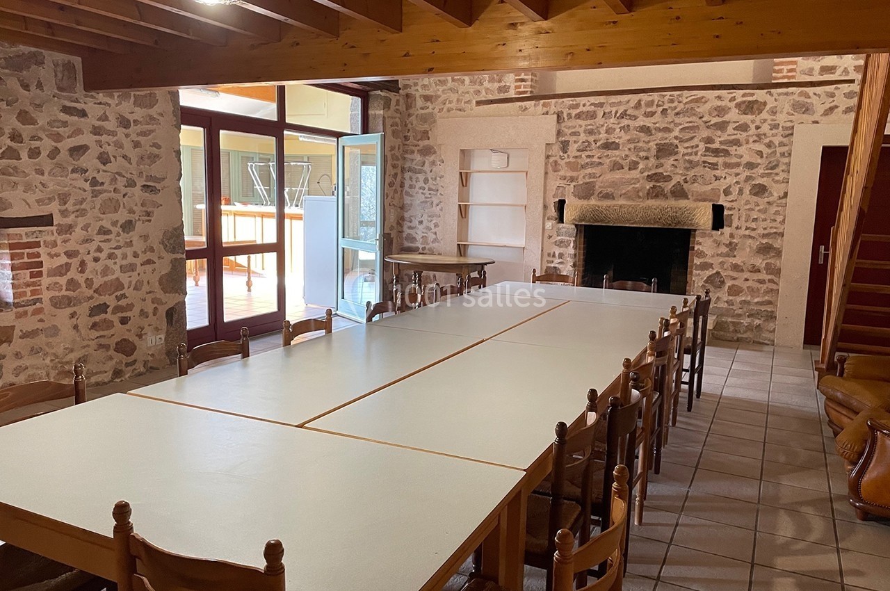 Salle avec des murs en pierre, une grande table entourée de chaises, une cheminée et une porte ouverte sur une terrasse.