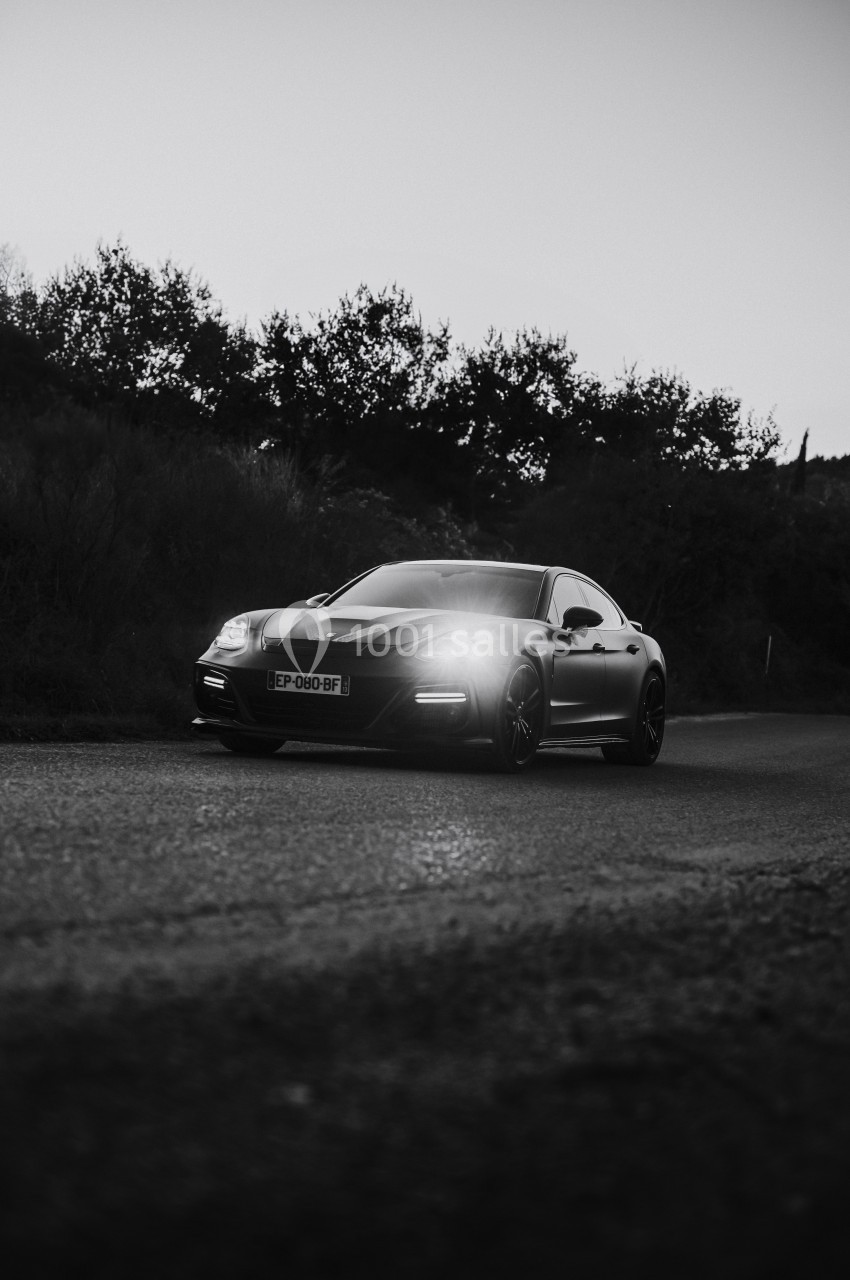 Voiture sportive noire stationnée sur une route, éclairée par ses phares, avec des arbres en arrière-plan.