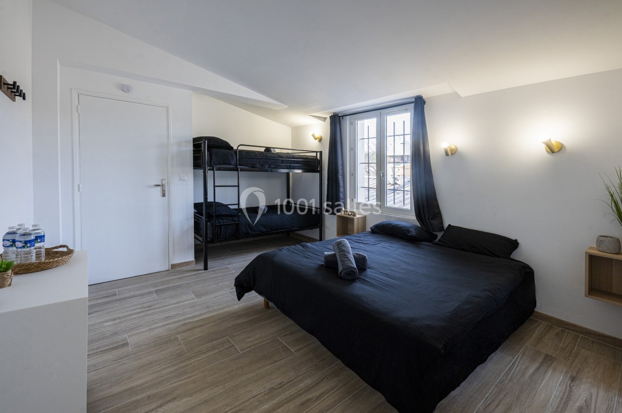 Chambre lumineuse avec un lit double, des lits superposés, parquet clair, murs blancs et une fenêtre avec rideaux.