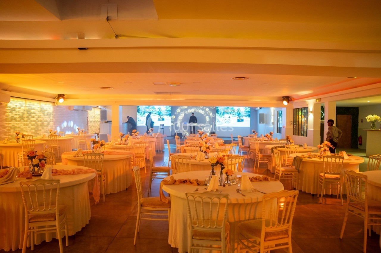 Salle de réception lumineuse avec tables rondes décorées de nappes et fleurs, chaises blanches et éclairage chaleureux.