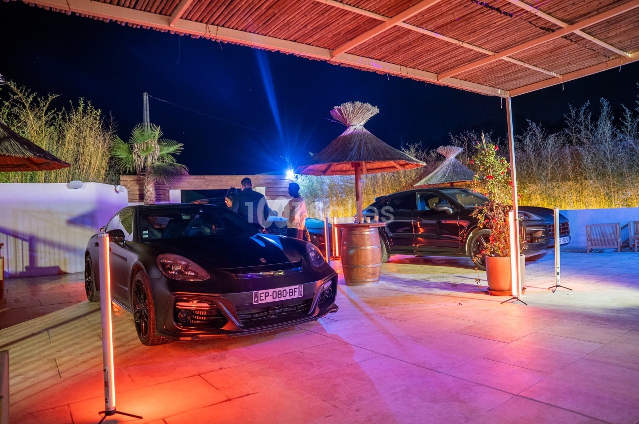 Deux voitures noires garées sur une terrasse éclairée par des lumières colorées, avec des parasols en arrière-plan.