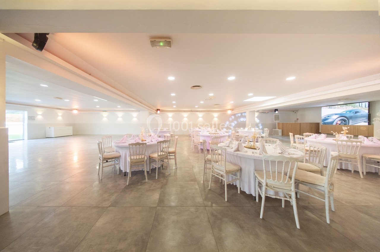 Salle de réception spacieuse avec tables rondes dressées, chaises blanches et éclairage doux.