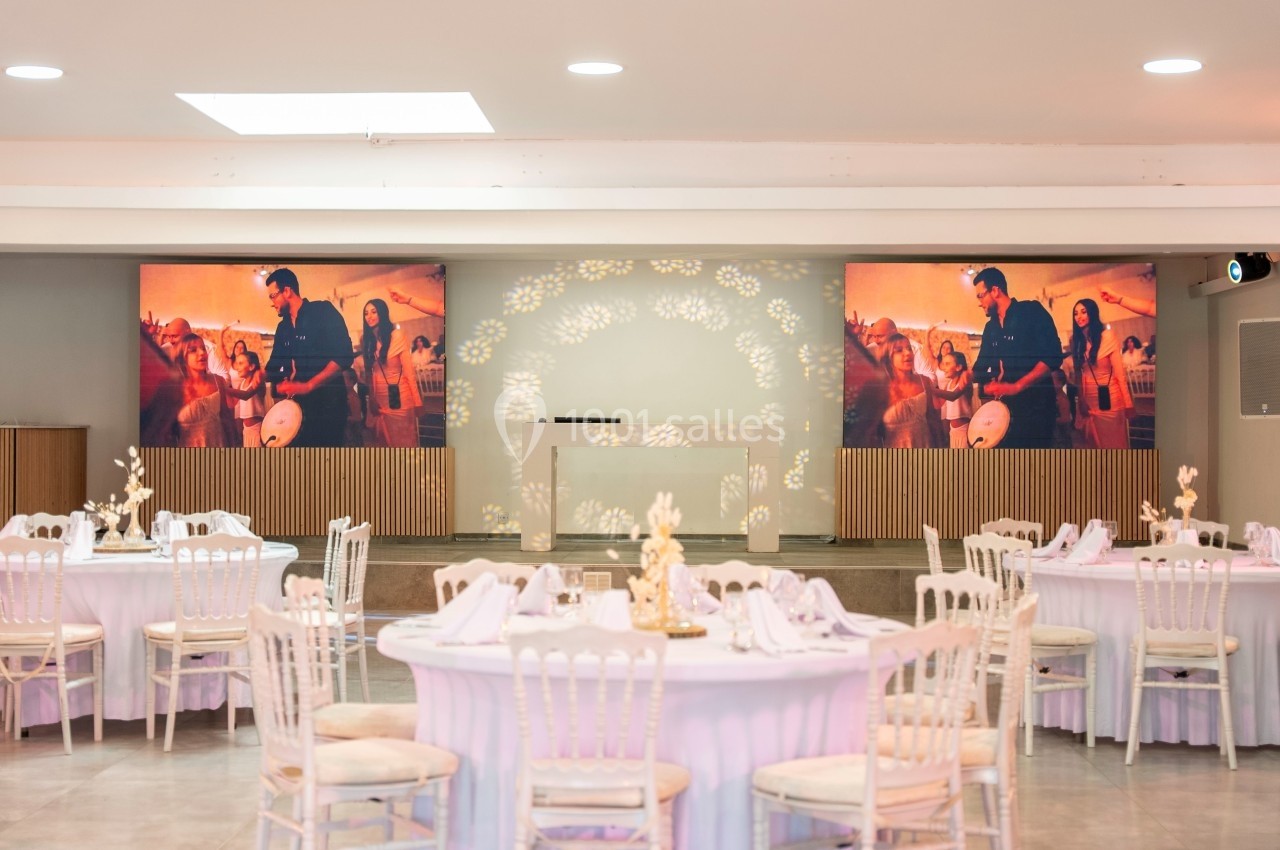 Salle de réception décorée avec des tables rondes élégantes et écrans affichant une scène festive en arrière-plan.