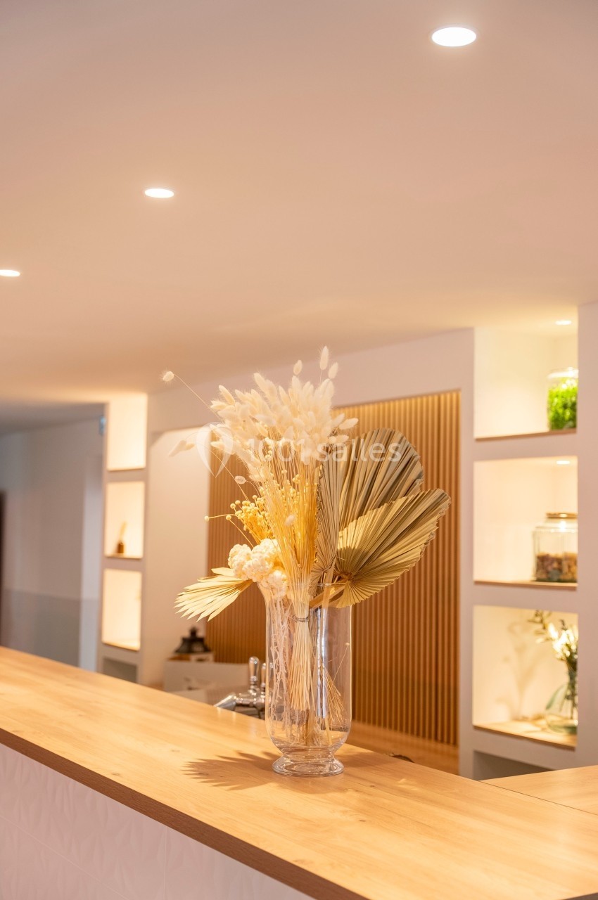 Vase en verre transparent contenant des fleurs séchées beige et dorées, posé sur un comptoir en bois clair.